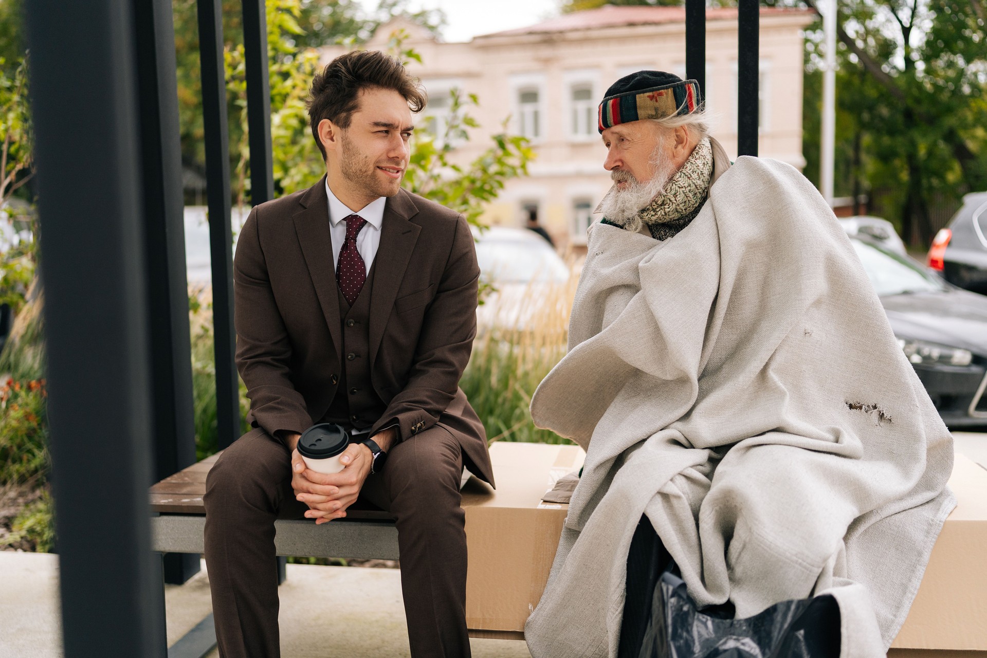 Empathetic businessman in suit listening story of old homeless man sitting on bench, representing growing gap between rich and poor in society.